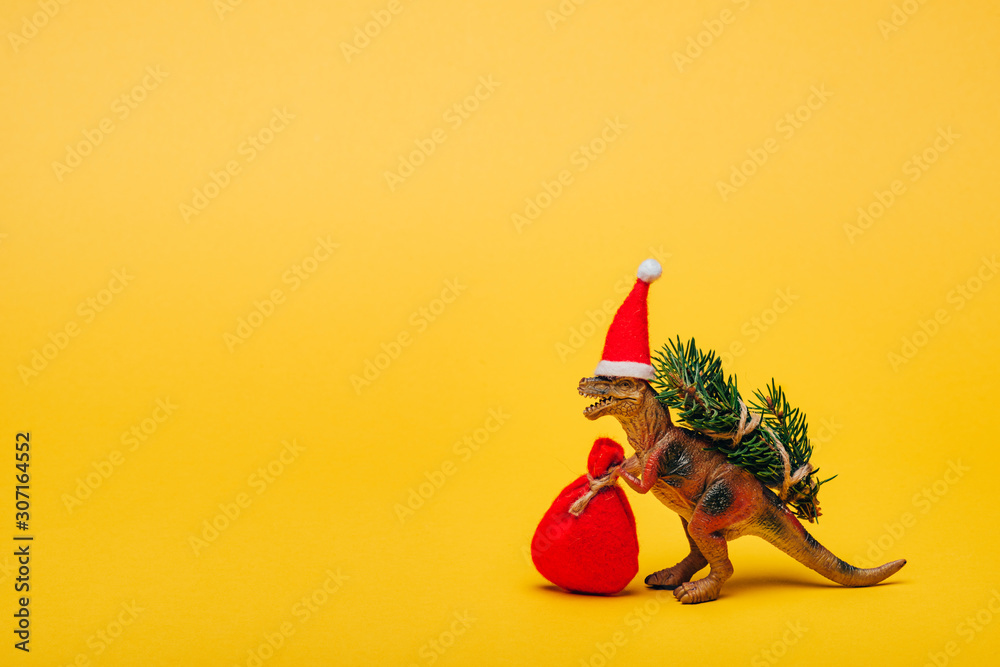 Fototapeta premium Zabawka dinozaur w santa hat z workiem i jodłą na żółtym tle
