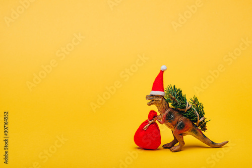 Fototapeta Naklejka Na Ścianę i Meble -  Toy dinosaur in santa hat with sack and fir on yellow background