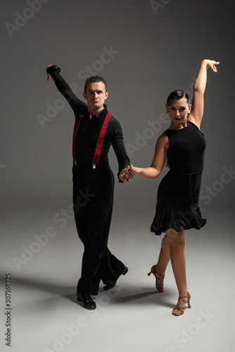 elegant dancers in black clothing looking at camera while dancing tango on grey background