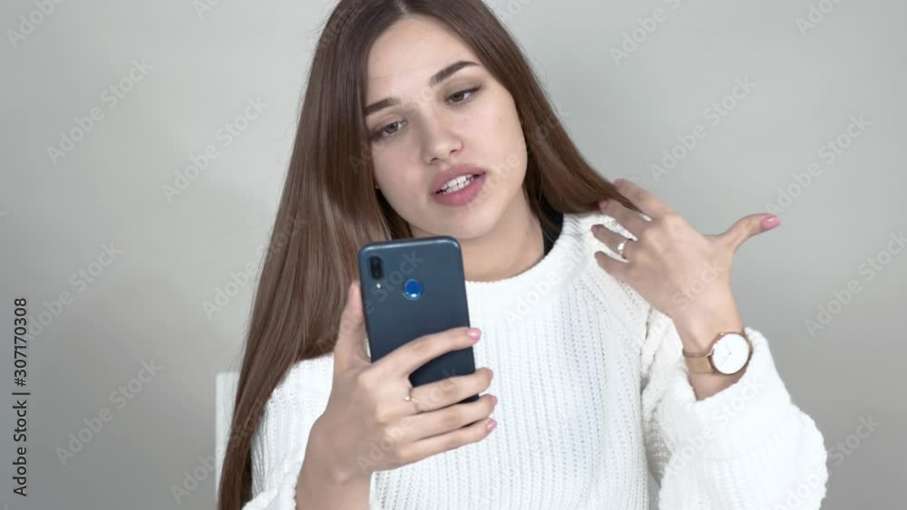 Gray background Caucasian girl white cardigan long straight brown-haired hair holds blue selfie communicates via video call. Close-up Gray background Caucasian girl white cardigan long straight brown-haired hair holds blue selfie communicates via video call. Close-up