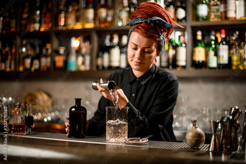 Bartender girl with a red hair pouring a alcoholic drink from the jigger to a measuring glass cup