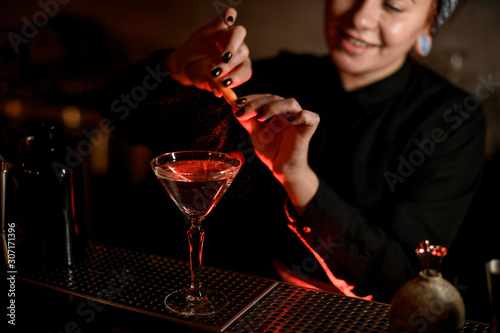 Smiling bartender girl sprinkling on a alcoholic drink in the cocktail glass with a orange zest juice