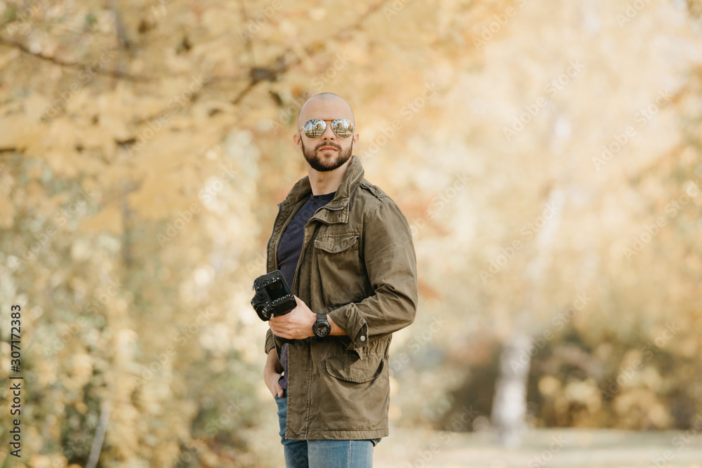 Bald photographer with a beard in aviator sunglasses with mirror lenses, olive cargo military jacket, blue shirt with digital wristwatch holds his camera and poses in the forest in the afternoon.