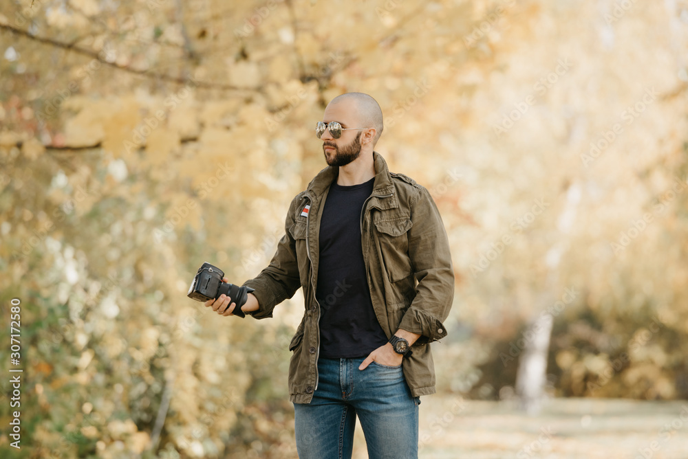 Bald photographer with a beard in aviator sunglasses with mirror lenses, olive cargo military jacket, blue jeans and shirt with digital wristwatch holds the camera by the lens and waits in the forest.
