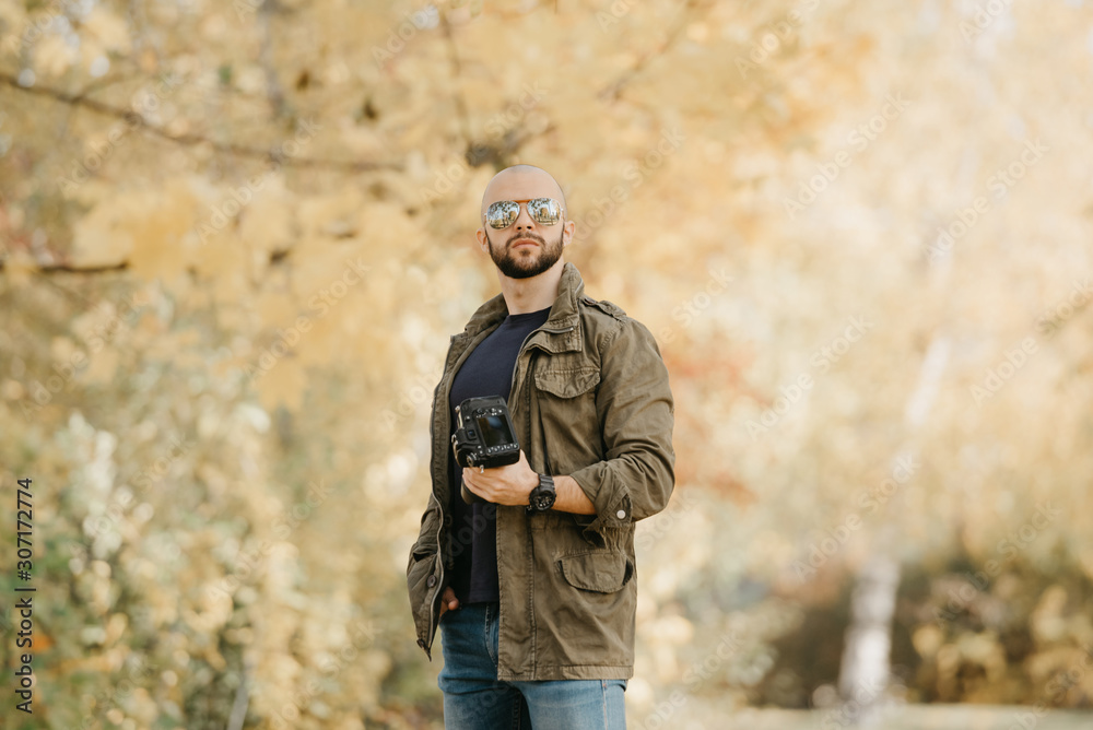 Bald photographer with a beard in aviator sunglasses with mirror lenses, olive military combat jacket, blue jeans and shirt with wristwatch poses holding his dslr camera and looks straight in the fore