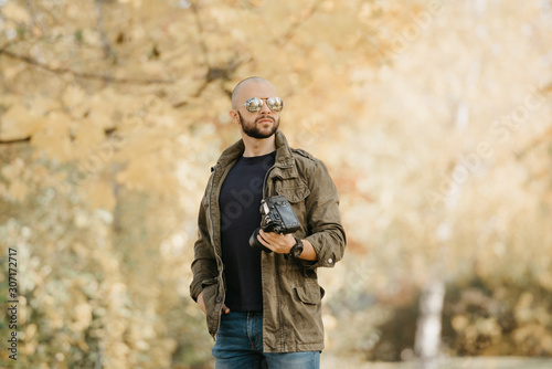 Bald photographer with a beard in aviator sunglasses with mirror lenses, olive military combat jacket, blue jeans and shirt with wristwatch poses with the camera and looks straight in the forest. © Roman Tyukin