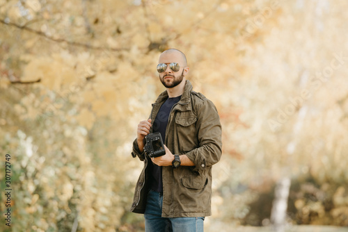 Bald photographer with a beard in aviator sunglasses with mirror lenses, olive military combat jacket, blue jeans and shirt with wristwatch poses holding the dslr camera and looks straight 