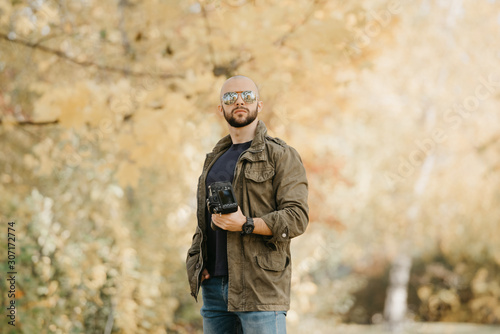 Bald photographer with a beard in aviator sunglasses with mirror lenses, olive military combat jacket, blue jeans and shirt with wristwatch poses holding his dslr camera and looks straight in the fore © Roman Tyukin