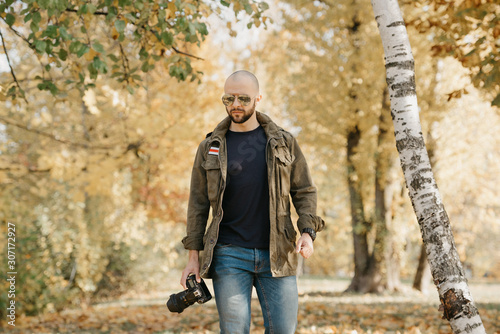 A bald journalist with a beard in aviator sunglasses with mirror lenses, olive military jacket, jeans and shirt with digital wristwatch holds the DSLR camera walks near the battlefield in the forest.