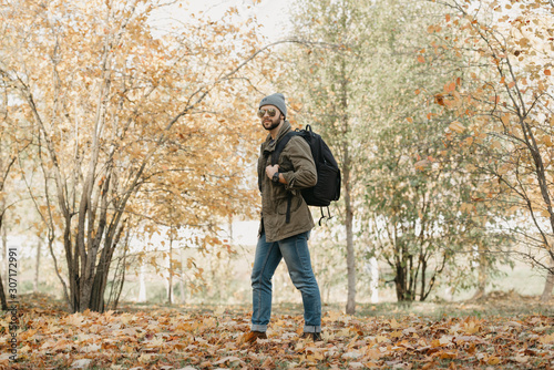 A brutal traveler with a beard in aviator sunglasses with mirror lenses, olive military combat jacket, jeans, hat with backpack and wristwatch walks in the afternoon in the forest. A photojournalist. © Roman Tyukin