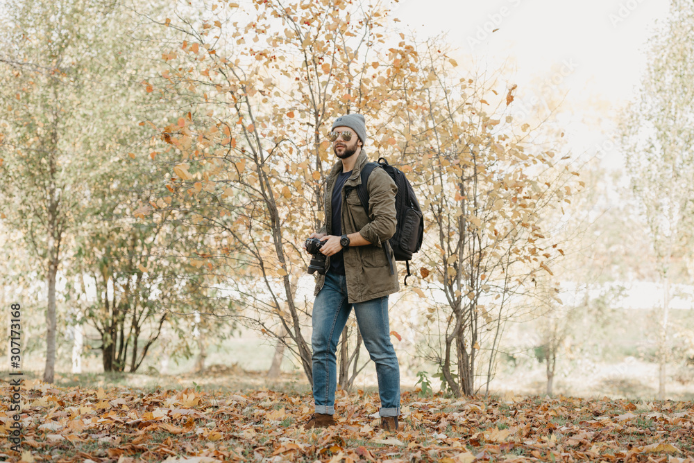 A photojournalist with a beard in aviator sunglasses with mirror lenses, olive military jacket, jeans, hat with backpack with wristwatch holds the DSLR camera, looks around near the battlefield A photojournalist with a beard in aviator sunglasses with mirror lenses, olive military jacket, jeans, hat with backpack with wristwatch holds the DSLR camera, looks around near the battlefield