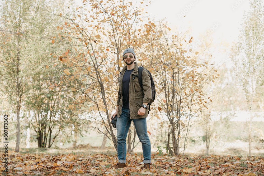 A military brave journalist with a beard in aviator sunglasses with mirror lenses, olive combat jacket, jeans and shirt with wristwatch holds the DSLR camera, looks around near the forest at the noon A military brave journalist with a beard in aviator sunglasses with mirror lenses, olive combat jacket, jeans and shirt with wristwatch holds the DSLR camera, looks around near the forest at the noon
