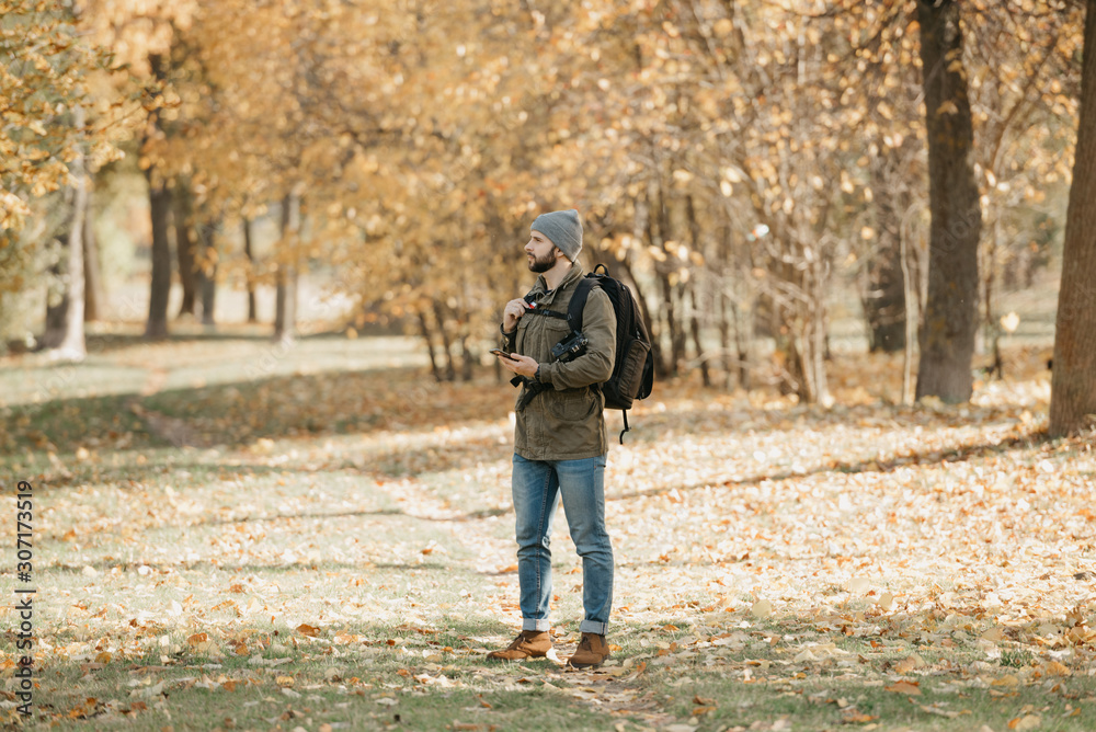 A traveler with a beard in olive military cargo combat jacket, jeans, hat with backpack and wristwatch holds the DSLR camera and the smartphone. A Photographer waits for someone at the noon. A traveler with a beard in olive military cargo combat jacket, jeans, hat with backpack and wristwatch holds the DSLR camera and the smartphone. A Photographer waits for someone at the noon.