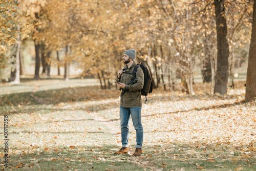 A traveler with a beard in olive military cargo combat jacket, jeans, hat with backpack and wristwatch holds the DSLR camera and the smartphone. A Photographer waits for someone at the noon.