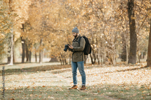 A traveler with a beard in olive military cargo combat jacket, jeans, hat with backpack and wristwatch holds the DSLR camera and checks something in the smartphone in the forest  at the noon. © Roman Tyukin