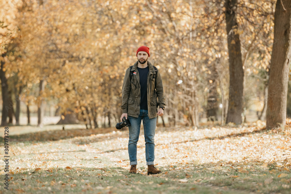 A photographer with a beard in olive military combat jacket, jeans, red hat with backpack and wristwatch holds the professional DSLR camera and waits in the forest at the afternoon A photographer with a beard in olive military combat jacket, jeans, red hat with backpack and wristwatch holds the professional DSLR camera and waits in the forest at the afternoon