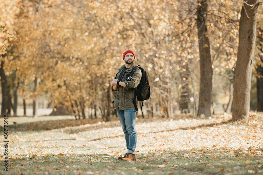 A brave photographer with a beard in olive military combat jacket, jeans, red hat with backpack holds the DSLR camera and his wristwatch in the forest at the afternoon A brave photographer with a beard in olive military combat jacket, jeans, red hat with backpack holds the DSLR camera and his wristwatch in the forest at the afternoon