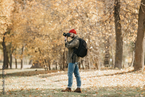 A photographer with a beard in an olive military cargo jacket, jeans, red hat with backpack and wristwatch takes photos with his professional DSLR camera in the forest at the noon © Roman Tyukin