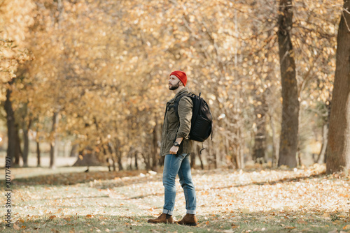 A brave traveler with a beard in an olive military combat jacket, jeans, red hat with a professional backpack and wristwatch walks in the afternoon in the forest. © Roman Tyukin