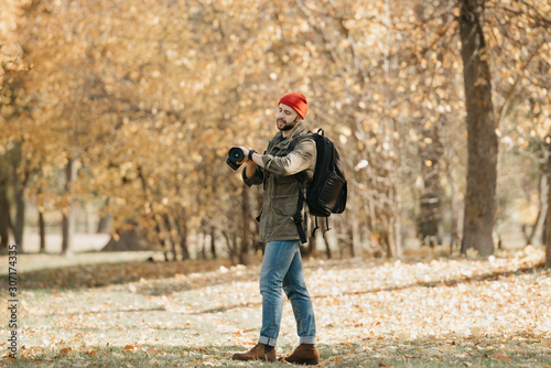 A photojournalist with a beard in an olive military cargo jacket, jeans, red hat with backpack holds his professional DSLR camera and checks the time on the wristwatch in the forest at the noon