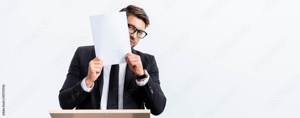 panoramic shot of scared businessman in suit standing at podium tribune and obscuring face with paper during conference isolated on white panoramic shot of scared businessman in suit standing at podium tribune and obscuring face with paper during conference isolated on white
