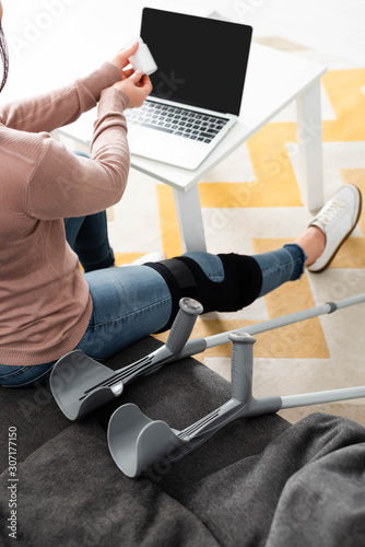 cropped view of woman with broken leg and crutches holding pills and having online consultation with doctor on laptop