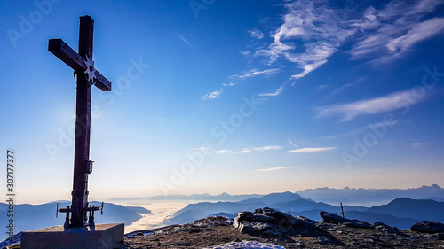 A metal cross on top of a mountain peak of Goldeck, Austria. The cross is towering above another points. In the back there are some snow caped mountain chains. Serenity and calmness.