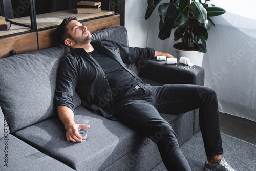 Quadro su tela high angle view of handsome man with panic attack holding glass of water and sit