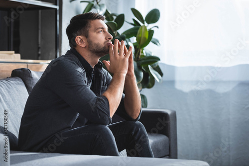 side view of handsome and pensive man sitting on sofa and looking up in apartment photo