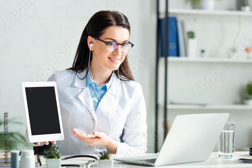 smiling doctor showing diagnosis on digital tablet while having online consultation on laptop