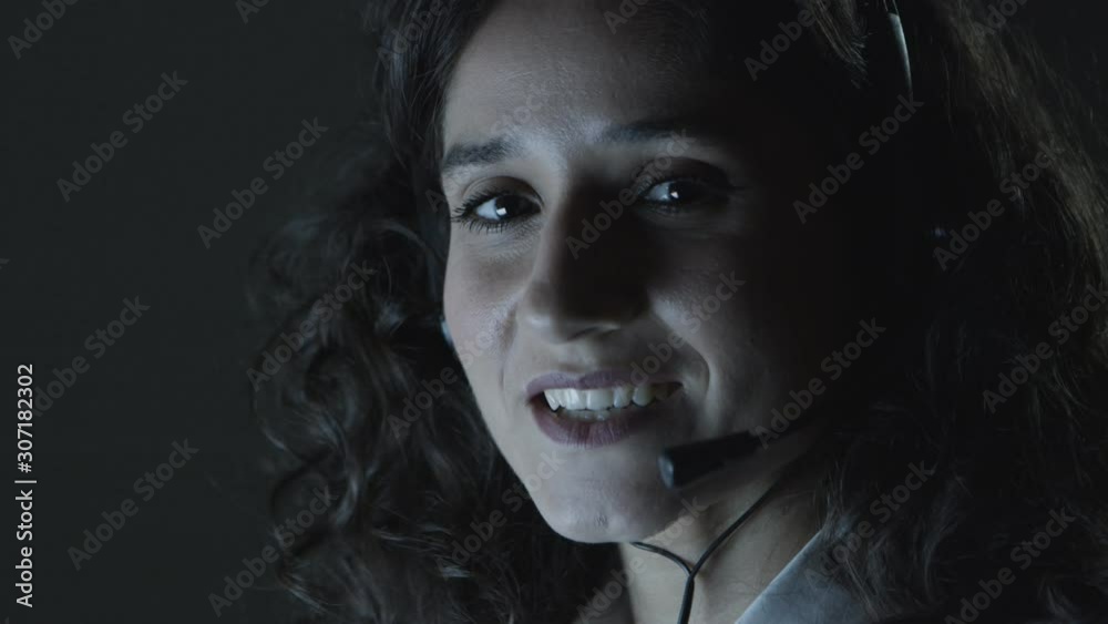 Happy woman in headset talking at camera. Close-up view of beautiful young female call center operator answering call and smiling at camera in dark office. Teleworking concept Happy woman in headset talking at camera. Close-up view of beautiful young female call center operator answering call and smiling at camera in dark office. Teleworking concept