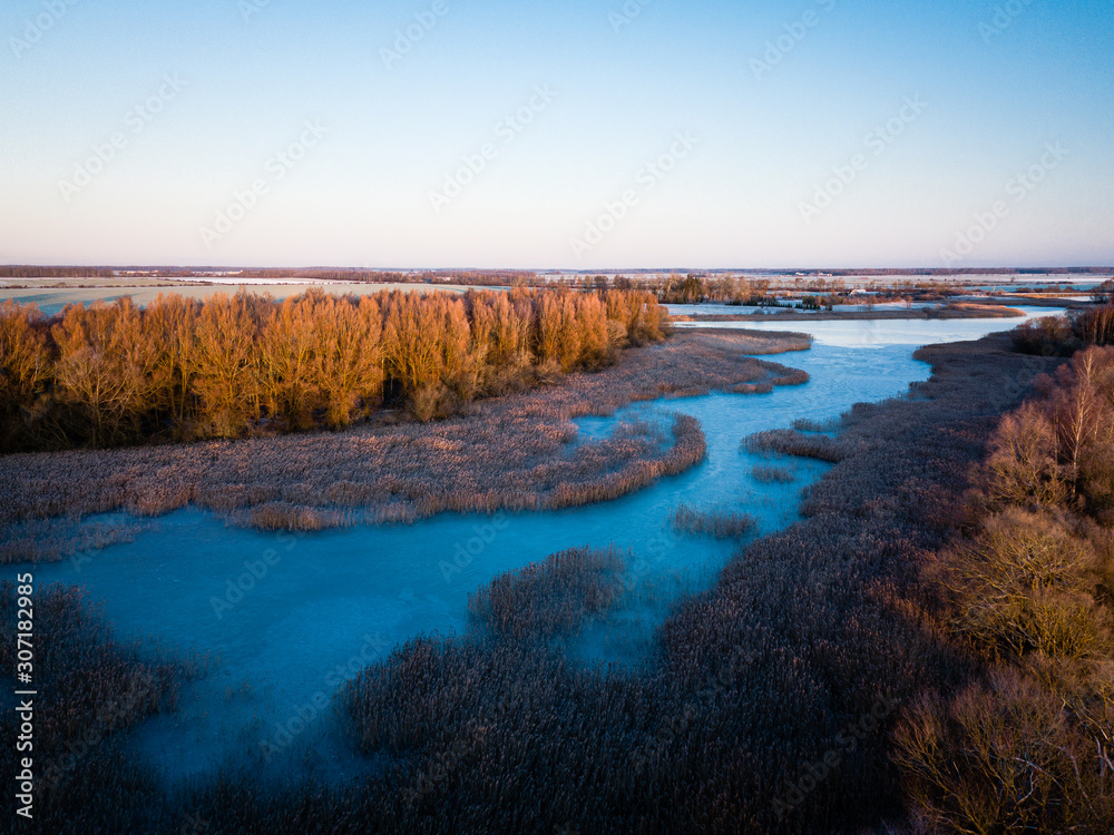 Aerial drone view of half frozen lake with island and trees - first sunrise light on tree tops - rural europe