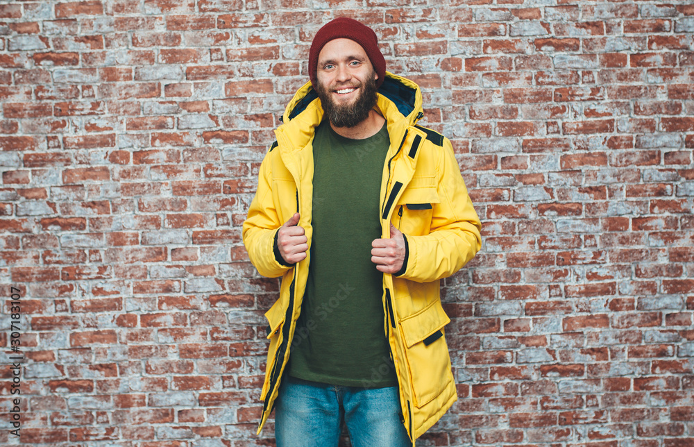 City portrait of handsome hipster guy with beard wearing a blank winter yellow jacket and red hat standing on a brick wall background. Empty space for your logo or design. Mockup for print. City portrait of handsome hipster guy with beard wearing a blank winter yellow jacket and red hat standing on a brick wall background. Empty space for your logo or design. Mockup for print.