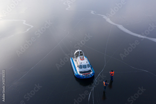 The river is covered with thin ice. On the icy surface is a hovercraft next to the boat are coast guard rescuers. The view from the top. photo