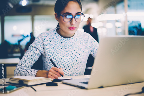 Serious female student in eyewear writing in netbook information from web page browsed on laptop computer,woman creating coursework project sitting at desktop with netbook for making research