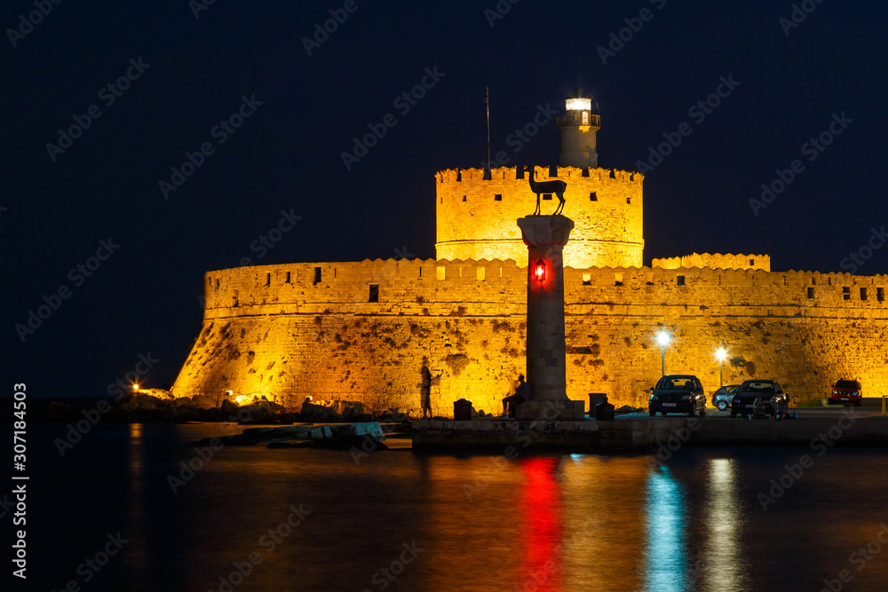 Night entrance to the port of Mandraki Rhodes island, illuminated ancient Fort, columns, reflection in the sea. Greece, Europe