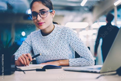 Portrait of skilled female employee in spectacles creating report sitting at desktop with modern laptop computer,pensive woman looking at camera analyzing information for startup at working place