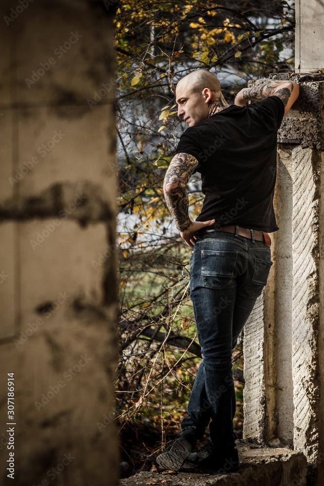 Bald brutal young man with tattoos stuffed on his arms and neck against the background of an abandoned destroyed building. Attractive tattooed stern guy. Concept of a bully and a dangerous guy.