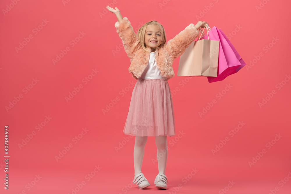 cute and smiling kid with outstretched hands holding shopping bags on pink background
