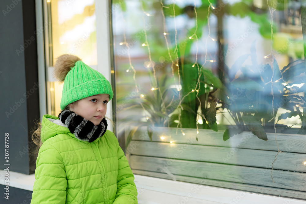 Young girl dressed in light green jacket and hat standing on the street at the glowing shop window.