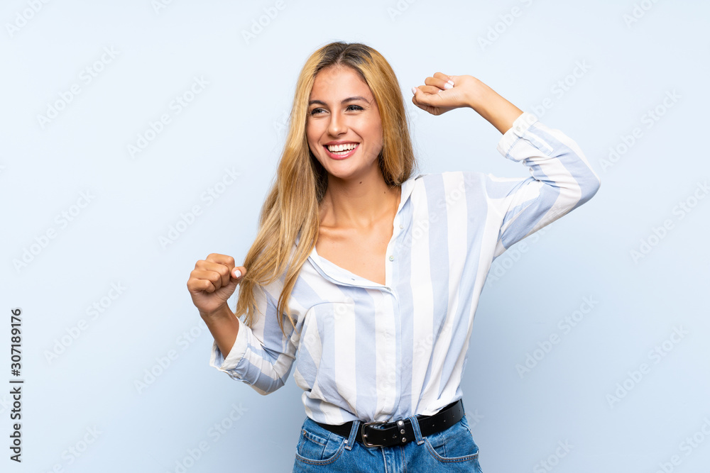 Young blonde woman over isolated blue background celebrating a victory - obrazy, fototapety, plakaty 