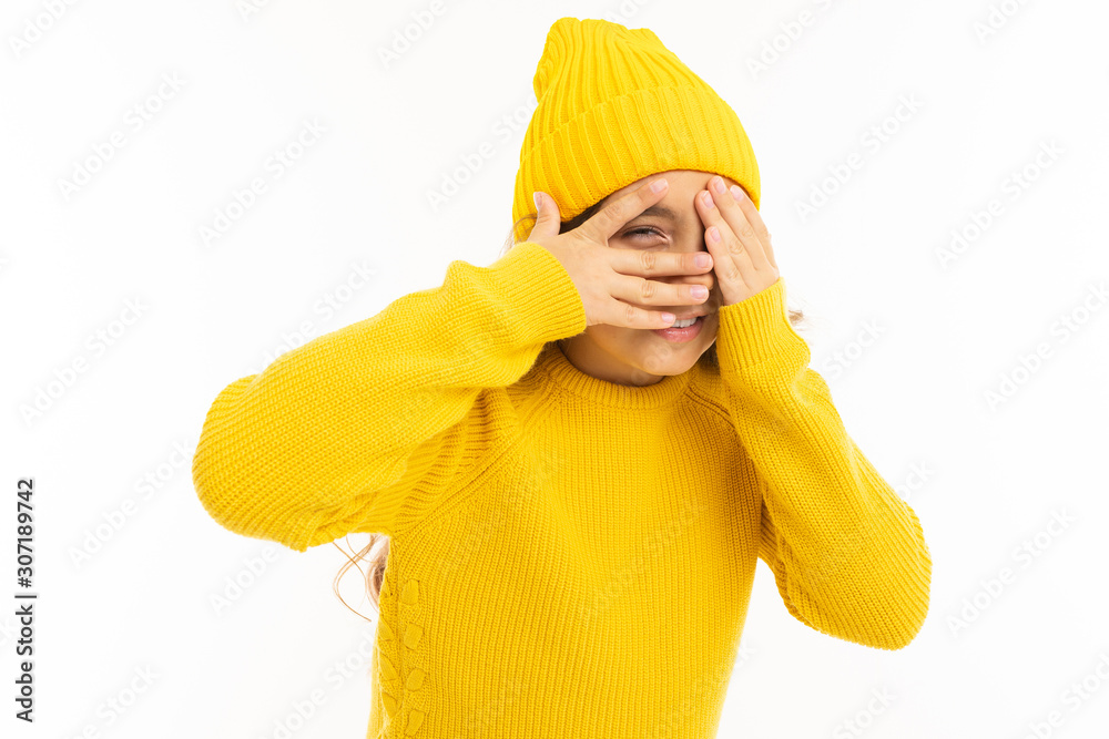 European attractive girl in a yellow hat and jacket covered her face with her hands and looks with one eye on a white studio background European attractive girl in a yellow hat and jacket covered her face with her hands and looks with one eye on a white studio background