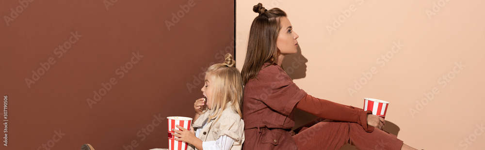 panoramic shot of daughter eating popcorn and mother on beige and brown background - obrazy, fototapety, plakaty 