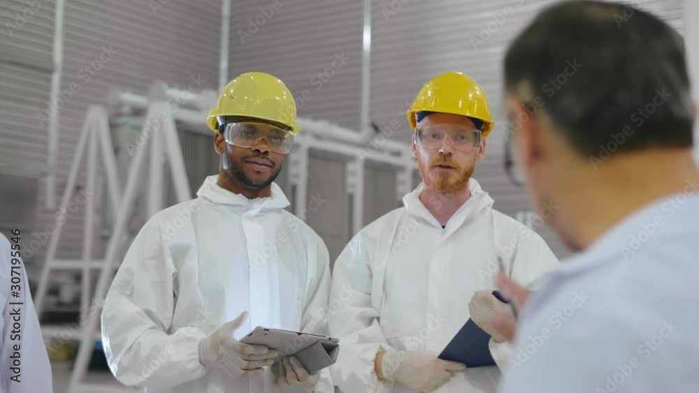 Engineer gives instructions to two workers in protective clothes and helmets. Multiracial group of scientists with tablet PC wearing white robes talk in research laboratory Engineer gives instructions to two workers in protective clothes and helmets. Multiracial group of scientists with tablet PC wearing white robes talk in research laboratory