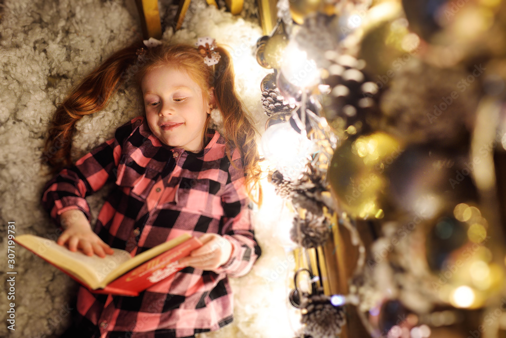 a little girl with red hair is lying under a Christmas tree, reading a book of fairy tales and smiling.