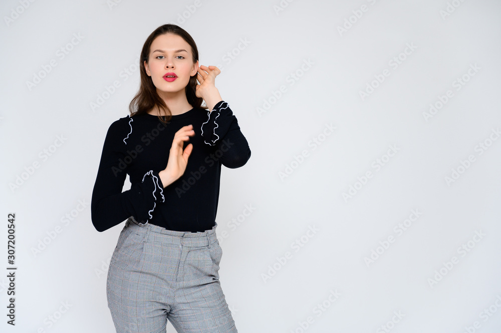 Concept adult girl on a white background. A photo of a pretty brunette girl in gray trousers and a black sweater smiles and shows different emotions in different poses right in front of the camera.