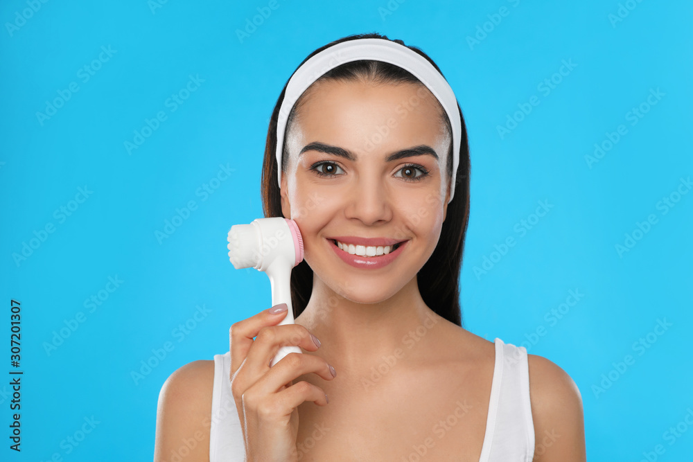 Young woman using facial cleansing brush on light blue background. Washing accessory