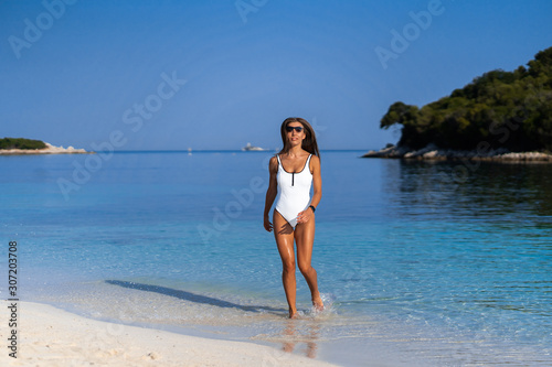 Fototapeta Naklejka Na Ścianę i Meble -  Young girl running on beach. Athletic woman jogging in trendy sexy white bodysuit enjoying the sun exercising. Healthy lifestyle. Fun walk along the shore. Perfect fitness body shapes and tan skin