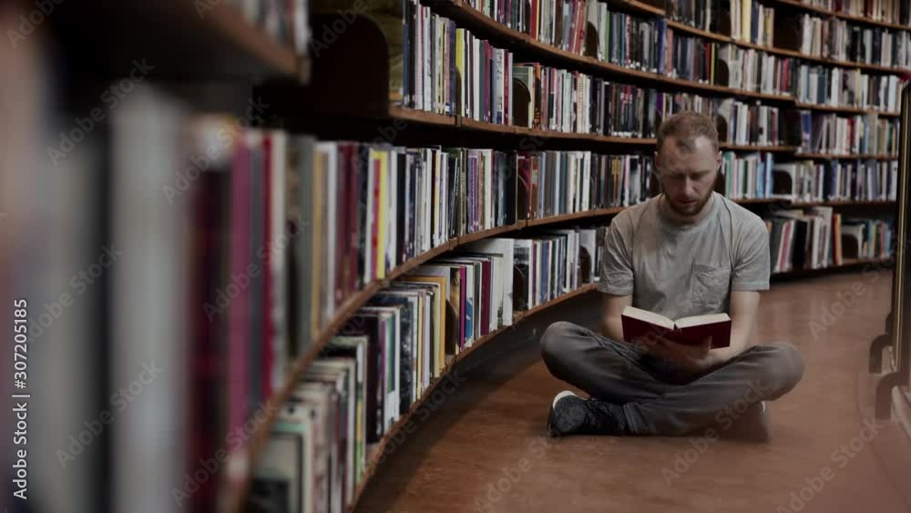 A man sits in a lotus position and reads a book among the shelves of the library. A man sits in a lotus position and reads a book among the shelves of the library.