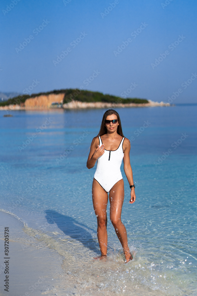 Young girl running on beach. Athletic woman jogging in trendy sexy white bodysuit enjoying the sun exercising. Healthy lifestyle. Fun walk along the shore. Perfect fitness body shapes and tan skin